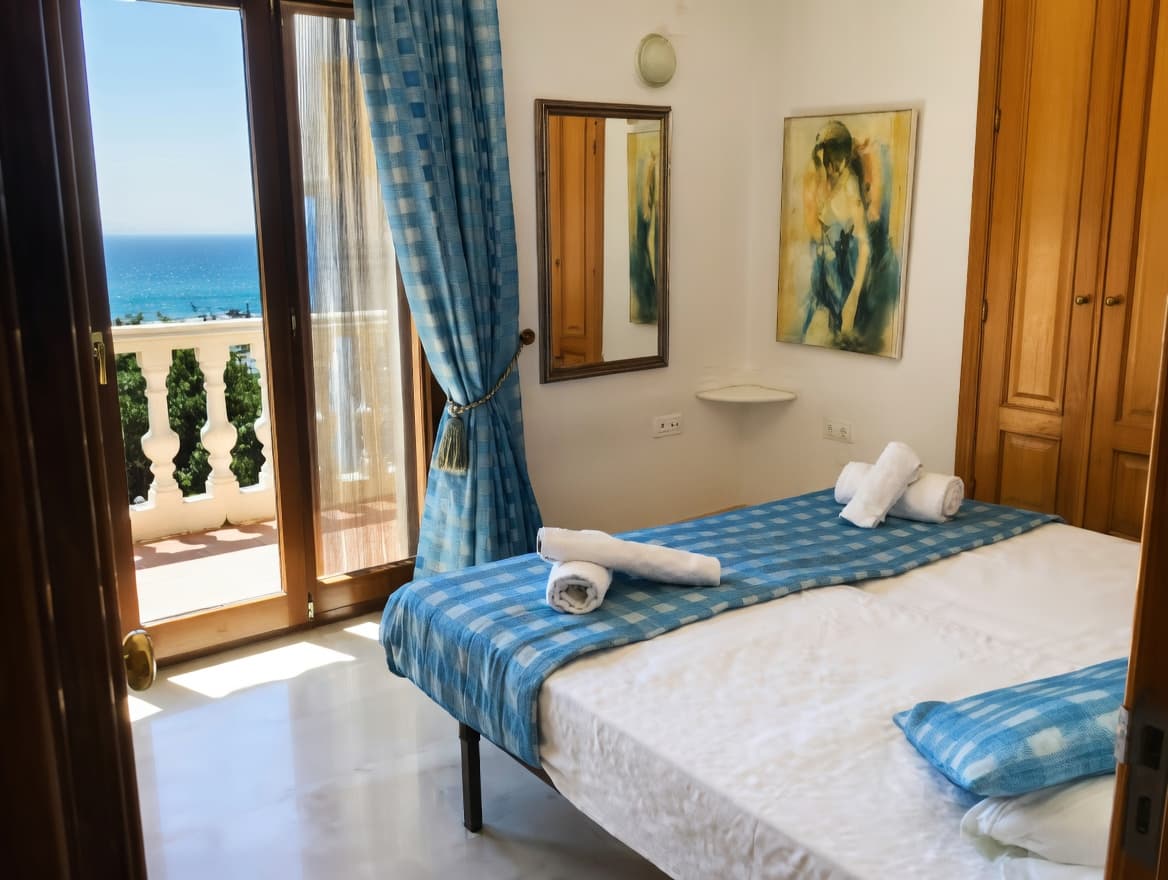 Dormitorio luminoso con cama, cortinas azules y balcón con vistas despejadas al mar azul.
