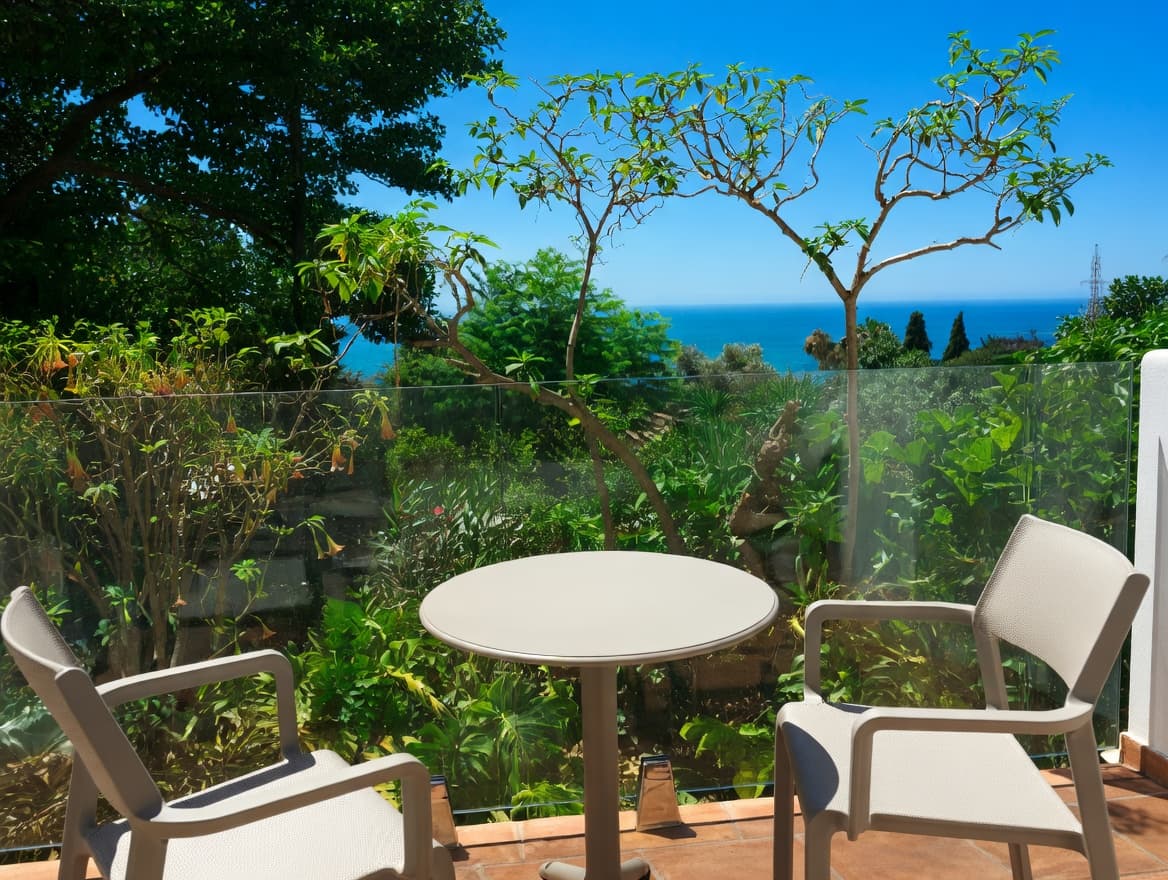 Terraza con mesa y sillas blancas, vistas al mar azul y vegetación tropical frondosa.