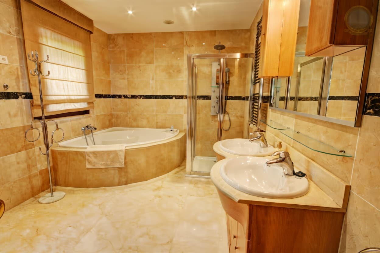 Baño lujoso con azulejos beige, bañera de esquina, ducha de cristal y lavabos dobles.