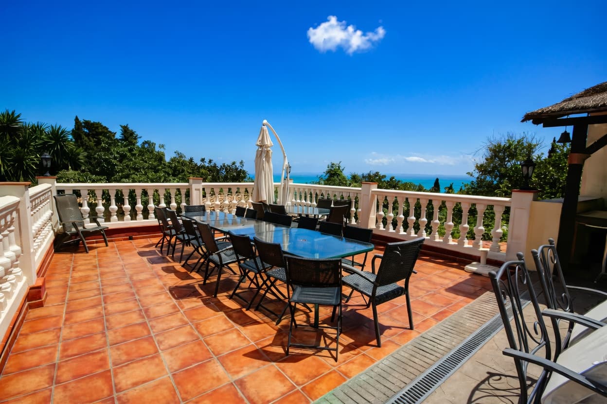 Terraza con mesa de cristal y sillas negras frente al mar bajo cielo azul.