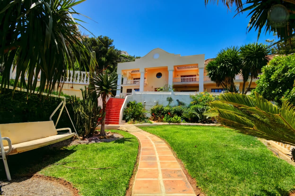 Villa color crema con jardín tropical, sendero de piedra, palmeras y un columpio blanco.