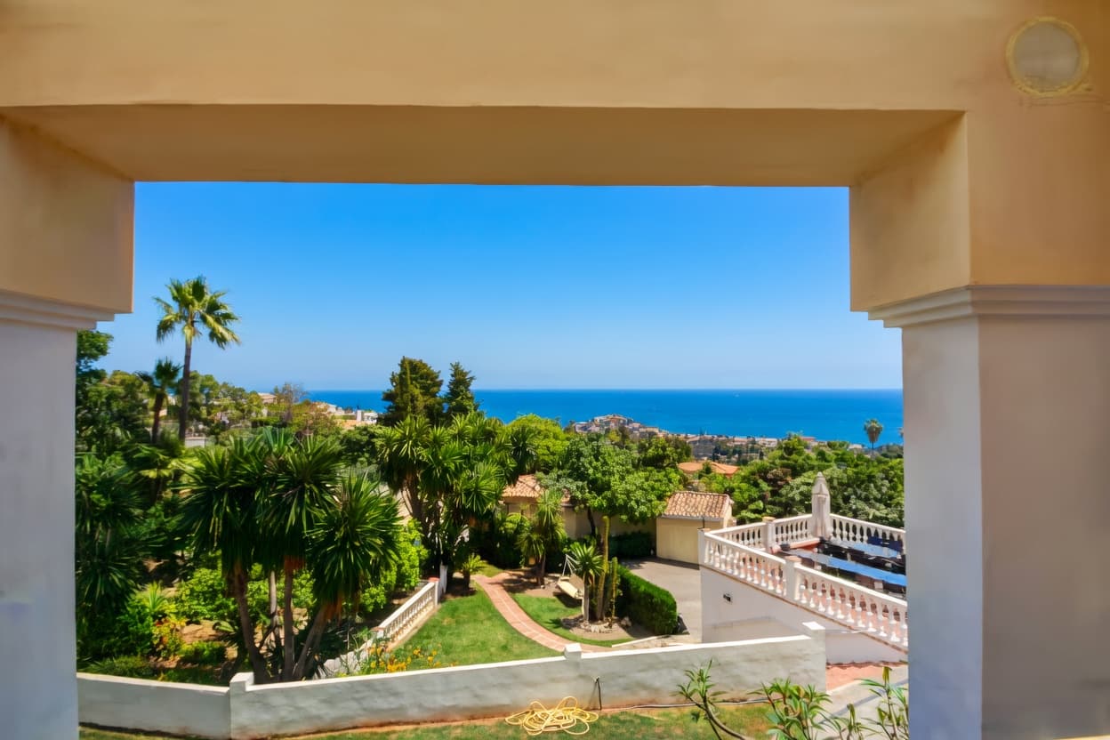 Vista enmarcada de un jardín tropical con palmeras frente al mar azul bajo cielo despejado.